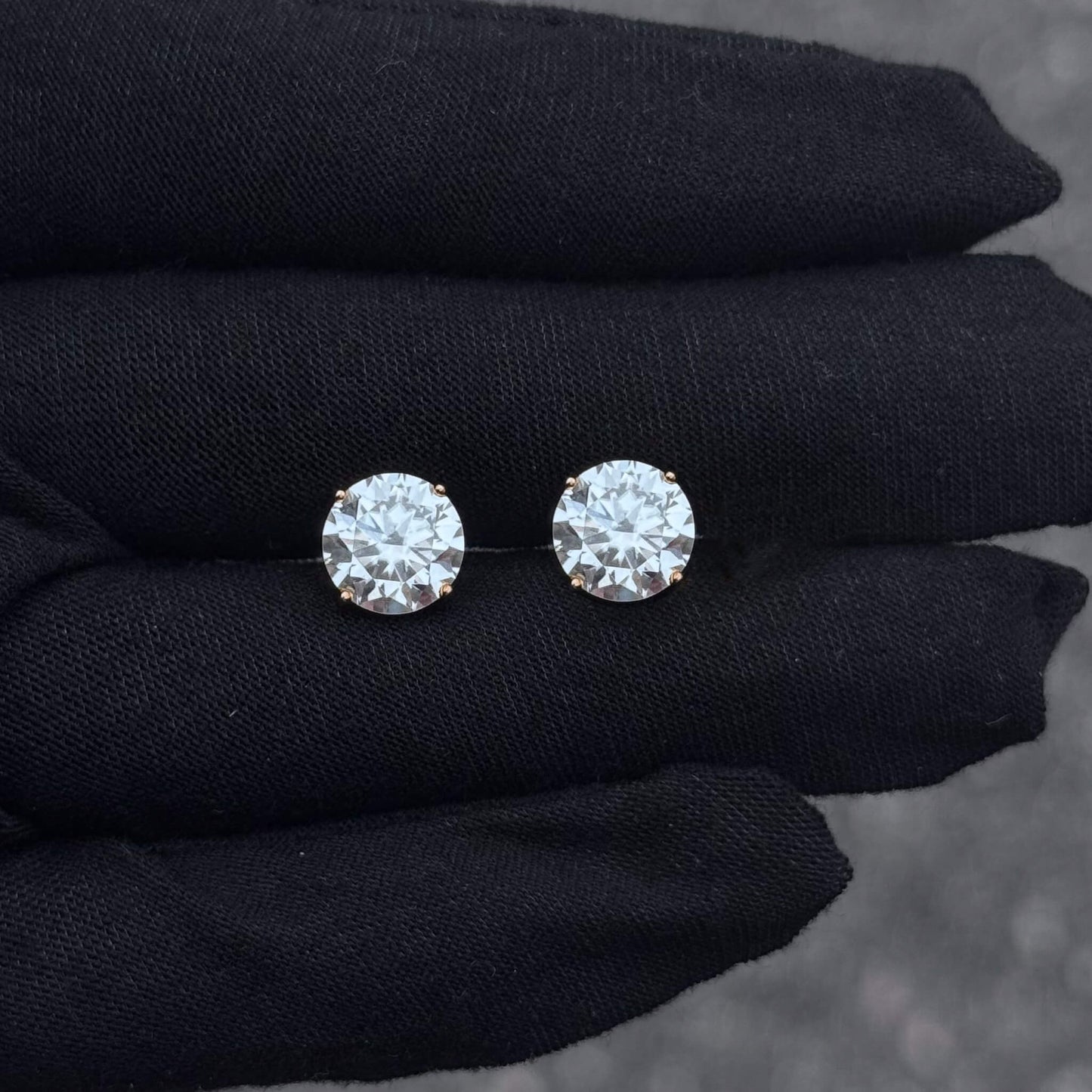 Pair of round moissanite solitaire stud earrings in sterling silver displayed on black glove.