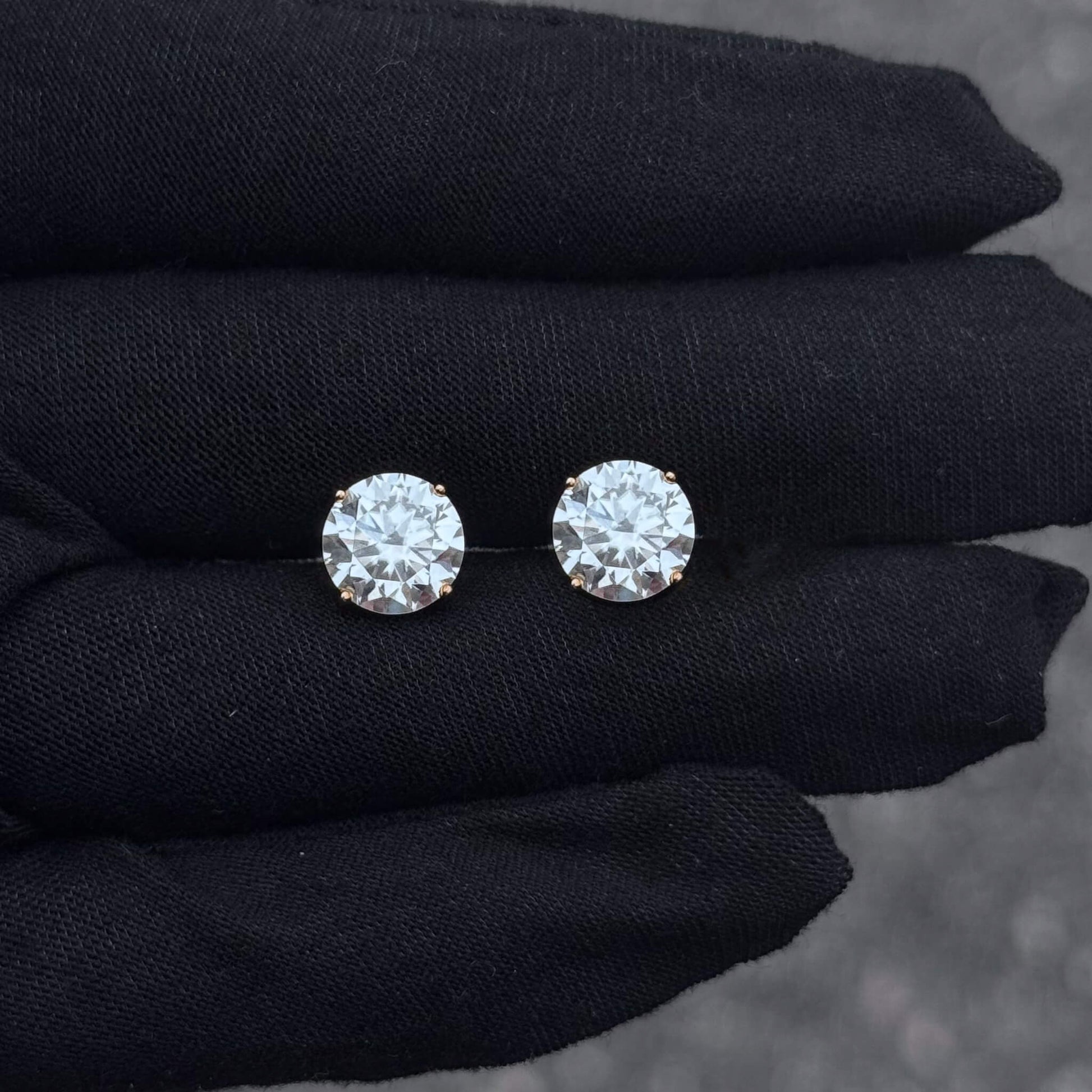 Pair of round moissanite solitaire stud earrings in sterling silver displayed on black glove.
